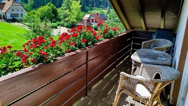 Un balcón con mobiliario de mimbre, baranda de madera y vista a un paisaje verde con casas.