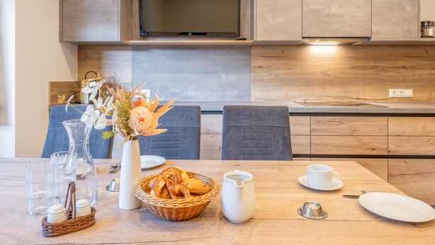 Una cocina moderna con armarios de madera, un televisor de pantalla plana y una mesa de comedor decorada con un florero, pan y una jarrilla.
