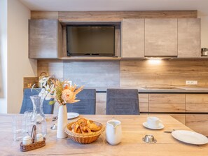 Eine moderne Küche mit Holzschränken, einem Flachbildfernseher und einem Esstisch, gedeckt mit einem Blumenstrauß, Brot und einer Kanne.