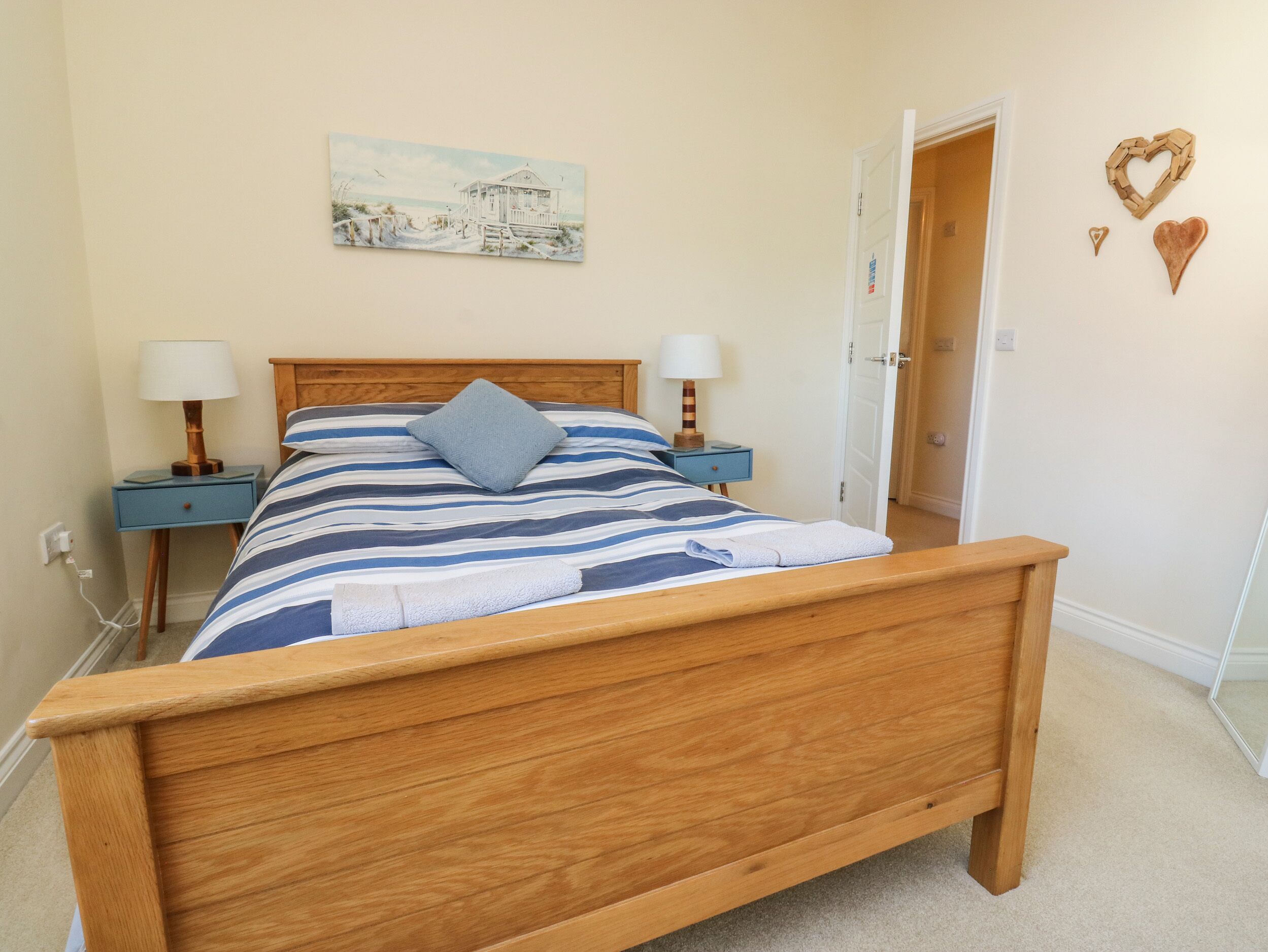 Un dormitorio con cama de madera, ropa de cama a rayas, dos mesitas de noche con lámparas, un cuadro en la pared y una puerta decorada con motivos de corazón.
