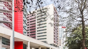 A modern building with a red facade and multiple floors, featuring a covered entrance area and a parking lot.