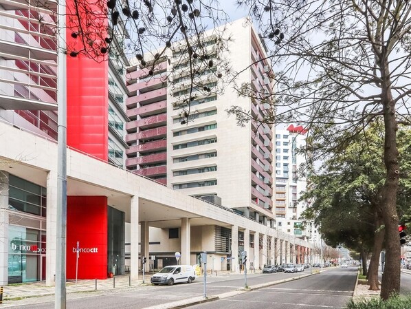 A modern building with a red facade and multiple floors, featuring a covered entrance area and a parking lot.