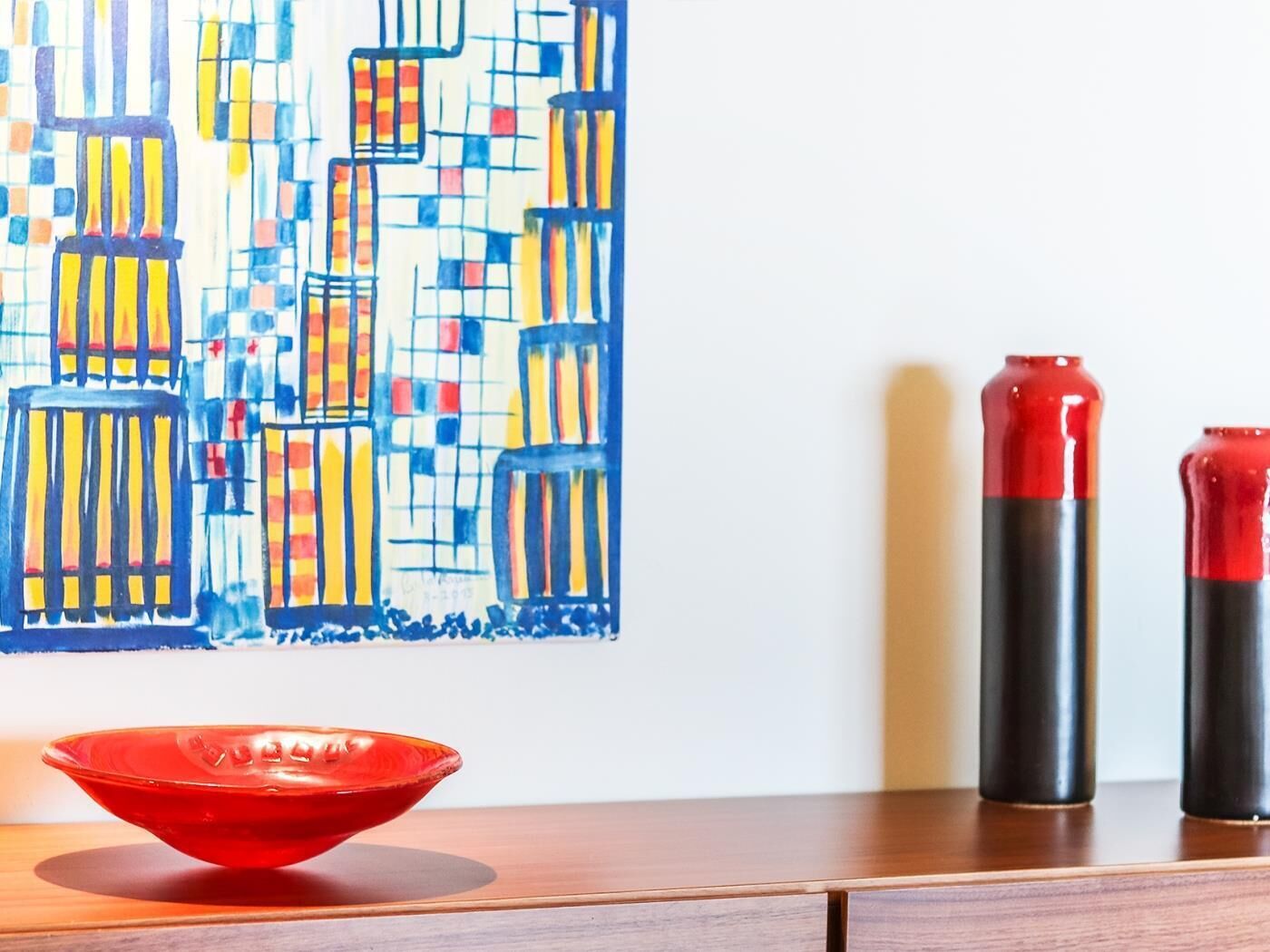 Uma mesa de madeira com uma tigela vermelha, duas velas pretas e vermelhas, e um quadro abstrato colorido na parede.
