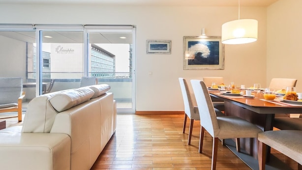 A modern hotel room with a sofa, dining table, chairs, and a view of a building through a glass door.