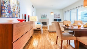 A modern living room with a wooden floor, a dining area with breakfast setup, and a large window offering a city view.