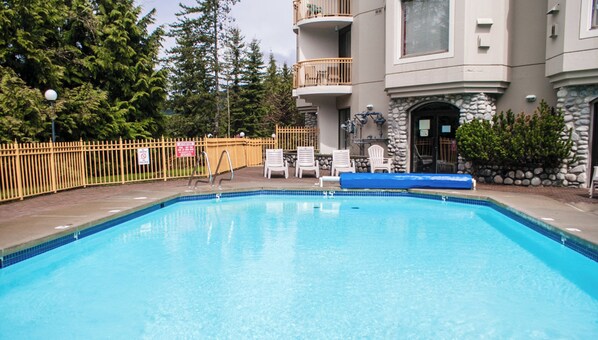 A rectangular swimming pool with a blue tiled edge, surrounded by a wooden fence. There are white lounge chairs and a blue floating lounge chair near the pool.