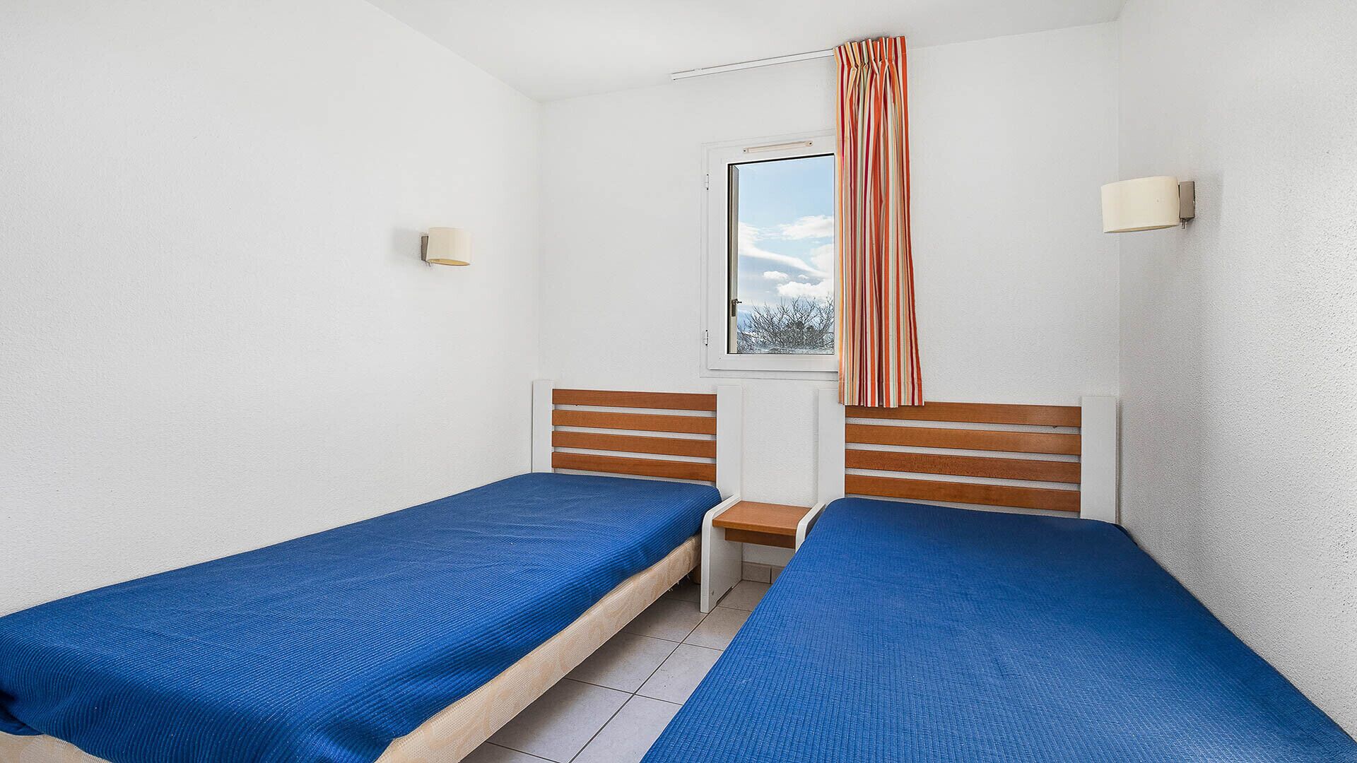 A small room with two single beds, a window with a view, a small table, and a wall-mounted lamp.