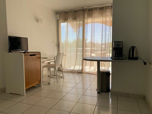 Una habitación con escritorio, silla, televisión, cafetera y un cubo de basura.