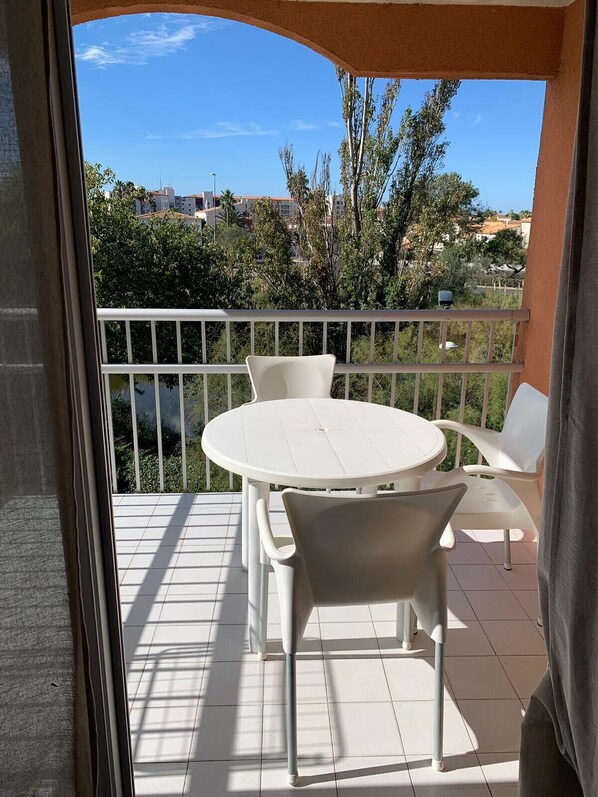 A balcony with a round table and chairs, a white railing, and a view of buildings and trees. - Apt 4 ppl near the sea (Agde)