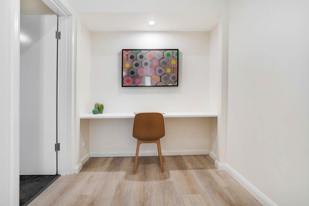 A hallway with a wooden floor, a white wall, and a framed artwork above a white shelf with a brown chair.