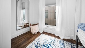 Ein Schlafzimmer mit großem Fenster, weißen Vorhängen, Holzboden, Spiegel, Bett und kleinem Teppich.