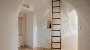 Ein schmaler Flur mit einer an der Wand angelehnten Holzleiter, einer Tür mit dekorativer Gestaltung und einem Regal mit einer Flasche.