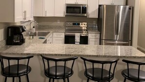 A kitchen with white cabinets, stainless steel appliances, a granite countertop, and four black bar stools.