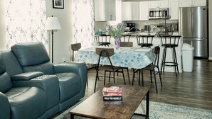 A modern living room with a blue sofa, a coffee table, and a dining area with a table and chairs. A kitchen with a refrigerator and microwave is visible in the background.