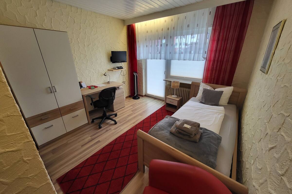 Ein kleines Hotelzimmer mit Bett, Schreibtisch, Stuhl, Kleiderschrank und einem Fenster mit Vorhängen.