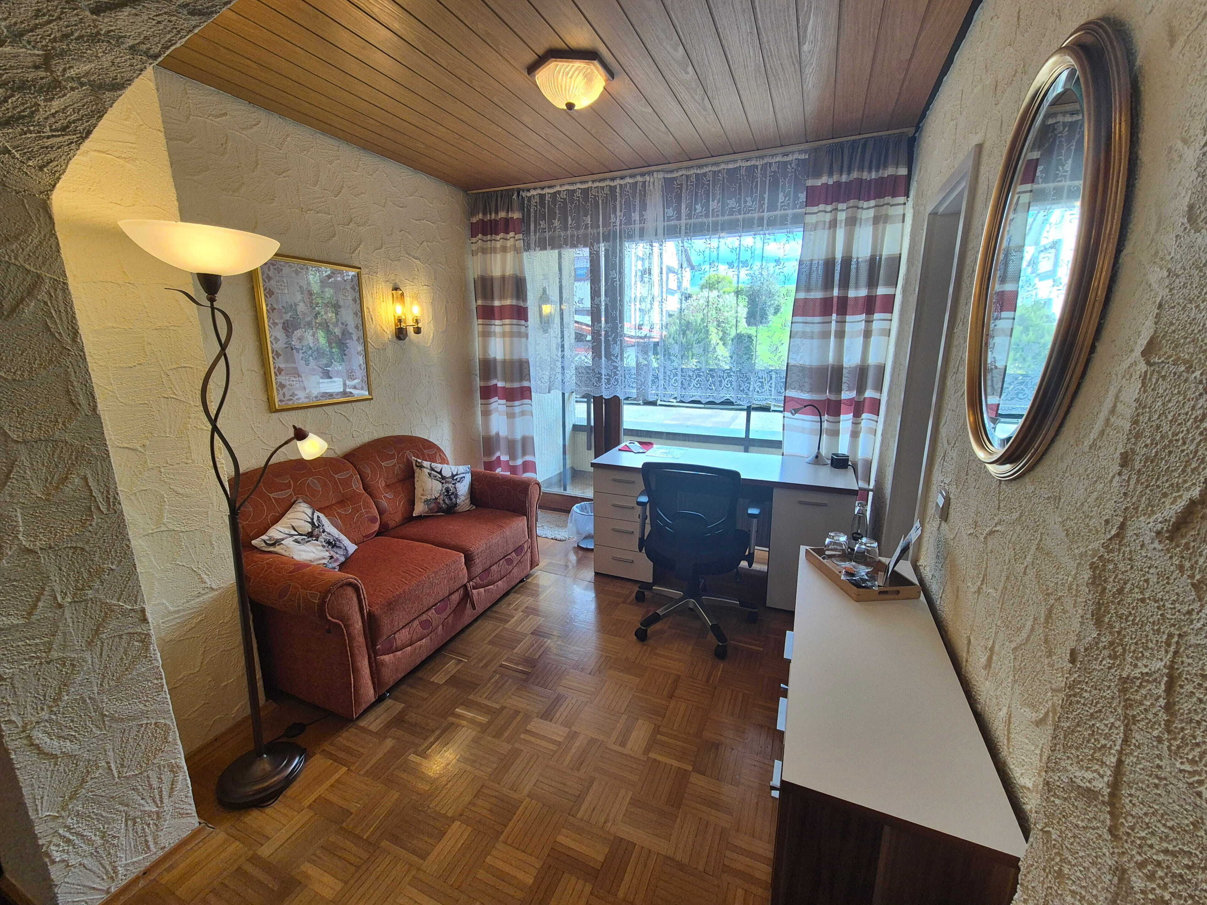 A room with a wooden ceiling, a sofa with patterned cushions, a desk with a chair, and a large mirror.