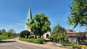 Een kerk met een hoge spits, een verharde weg en een oprit naar een gebouw met een pannendak.