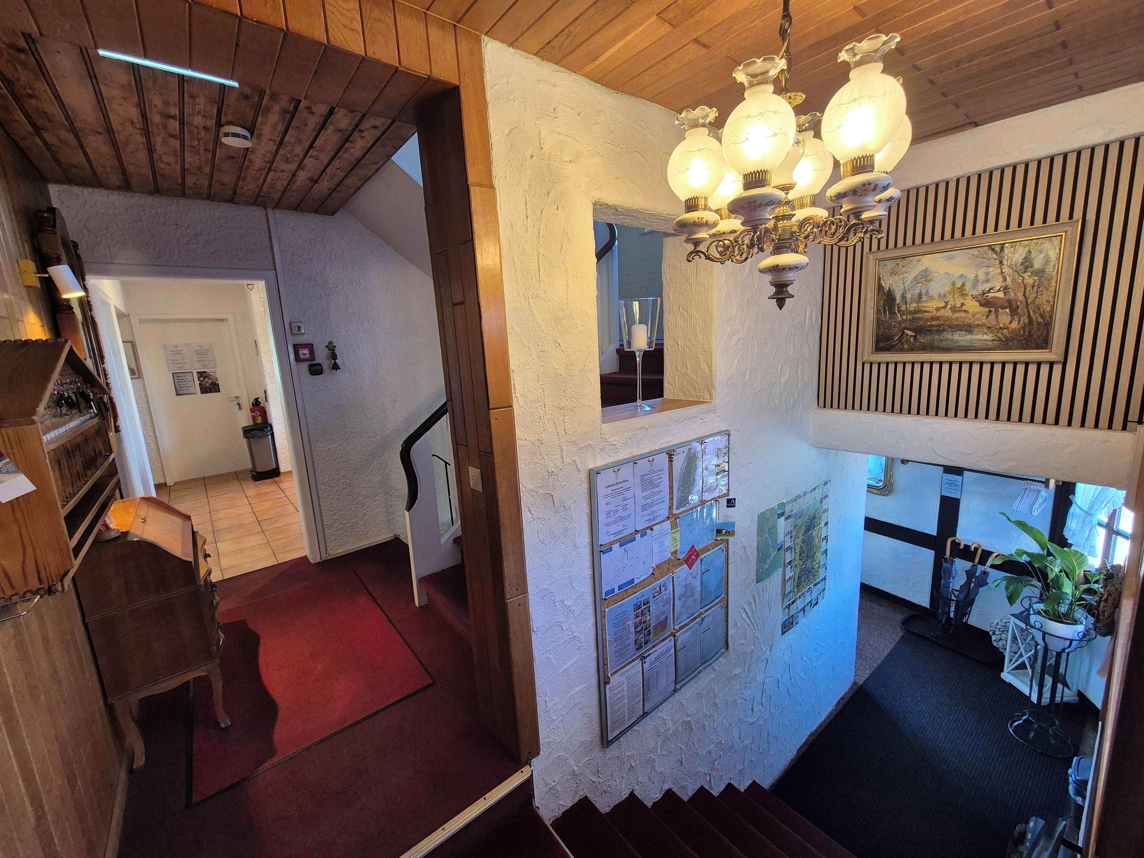 A wooden interior with a chandelier, a staircase, and a bulletin board with notices.