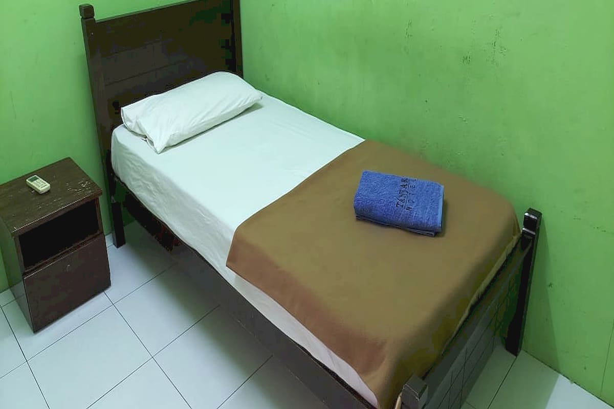 A single bed with a white pillow and a brown blanket, a small wooden nightstand with a remote control, and a blue towel on the bed.