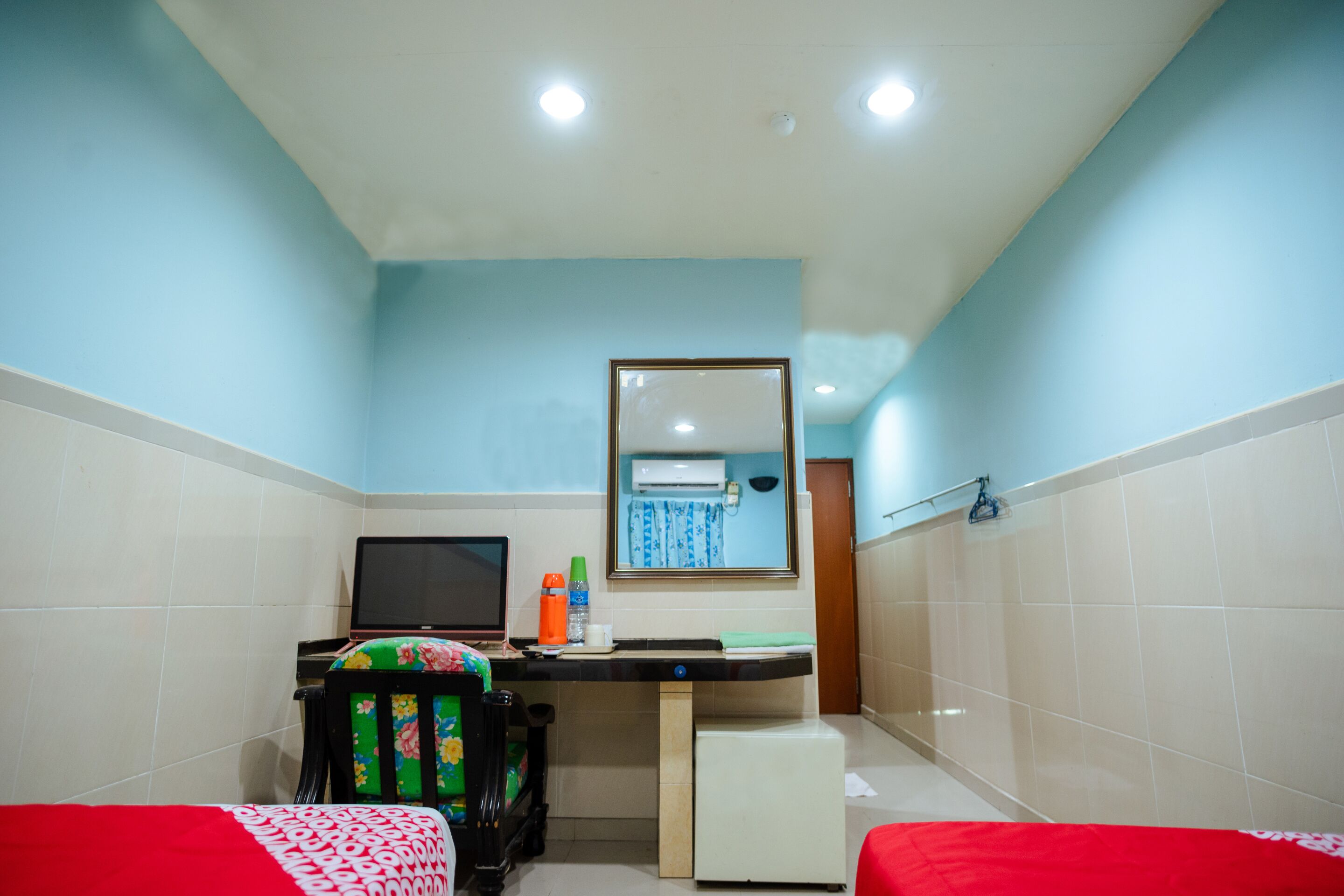 A small, brightly lit room with a blue wall, a white ceiling, and a tiled floor. It includes a desk with a computer, a chair, a small refrigerator, and a bed with a red and white bedspread.