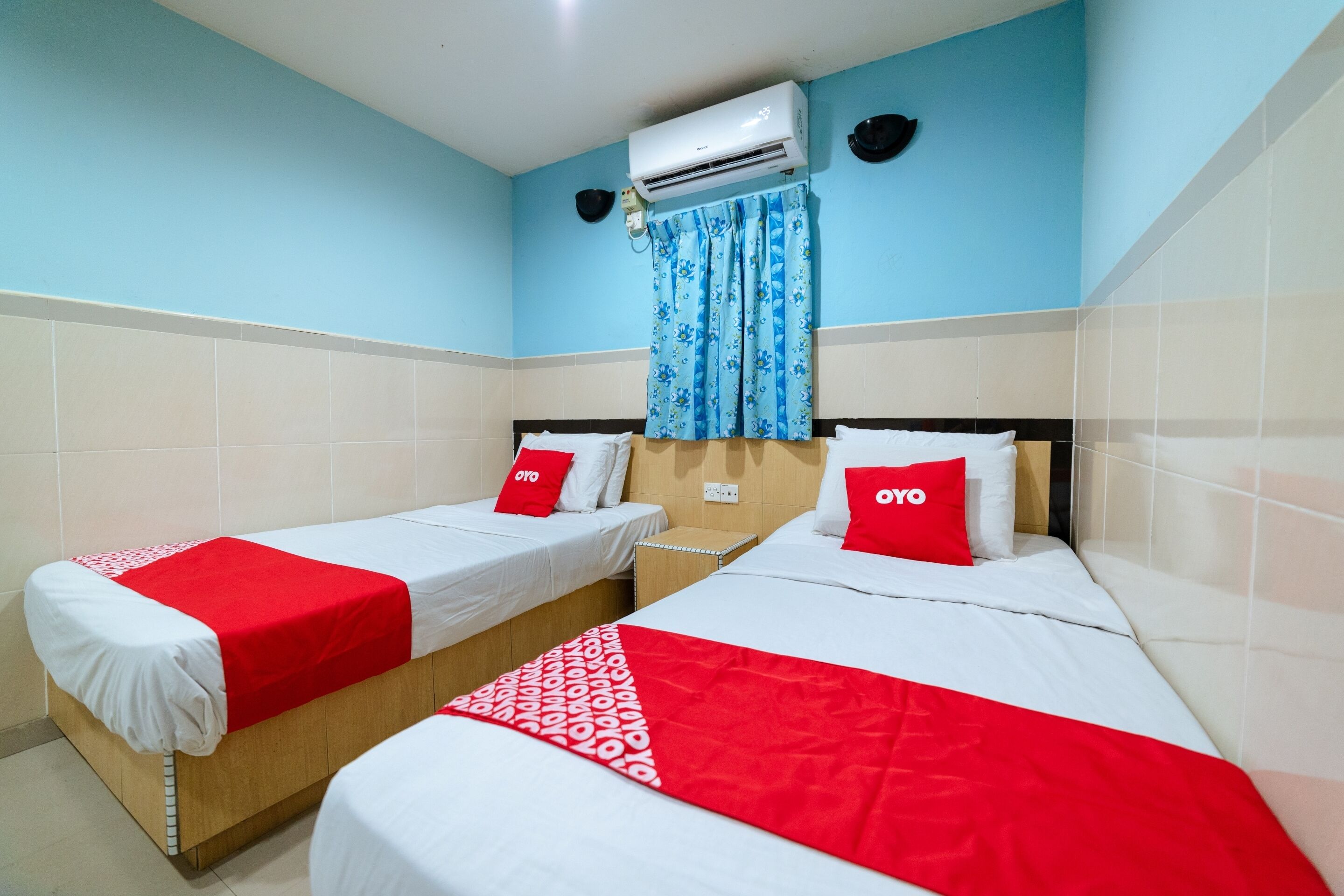Habitación de hotel con dos camas individuales, cada una con cubrecama y almohadas rojo y blanco. Unidad de aire acondicionado montada en la pared sobre las camas, y una ventana con cortina floral azul.