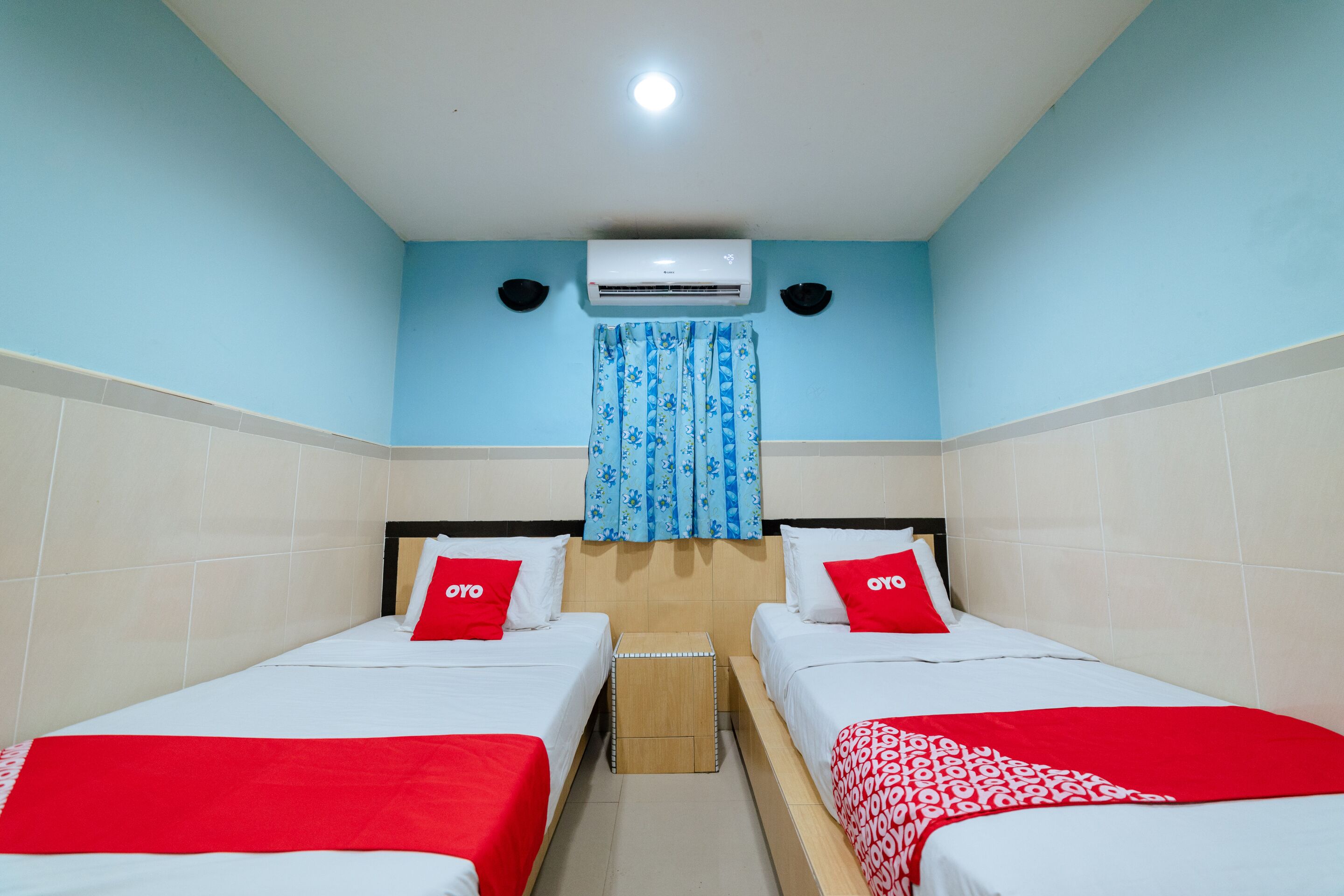 Habitación de hotel con dos camas, cada una con cubrecama rojo y blanco, y un taburete de madera pequeño entre ellas.