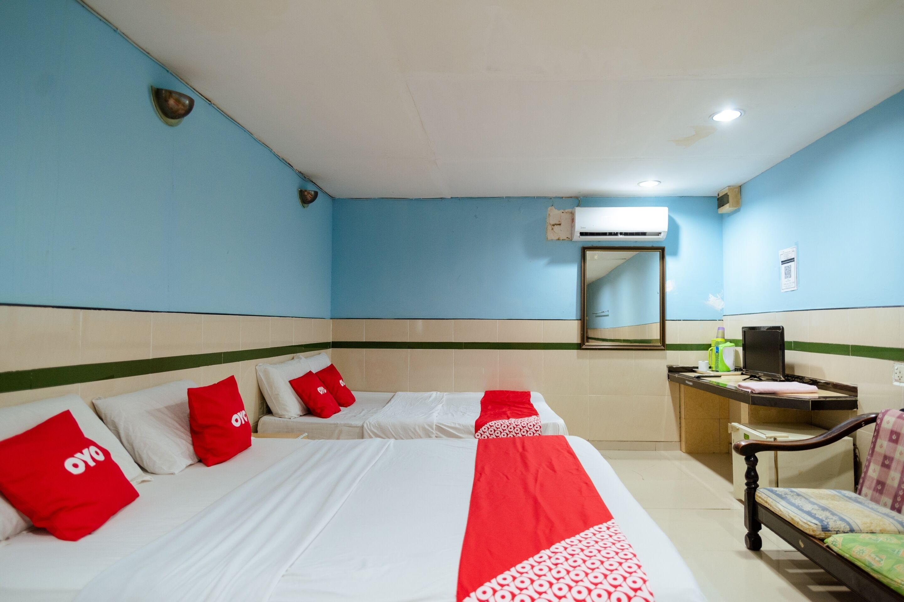 Quarto de hotel com duas camas, cada uma com roupa de cama vermelha e branca, uma escrivaninha com cadeira e um ar condicionado na parede.