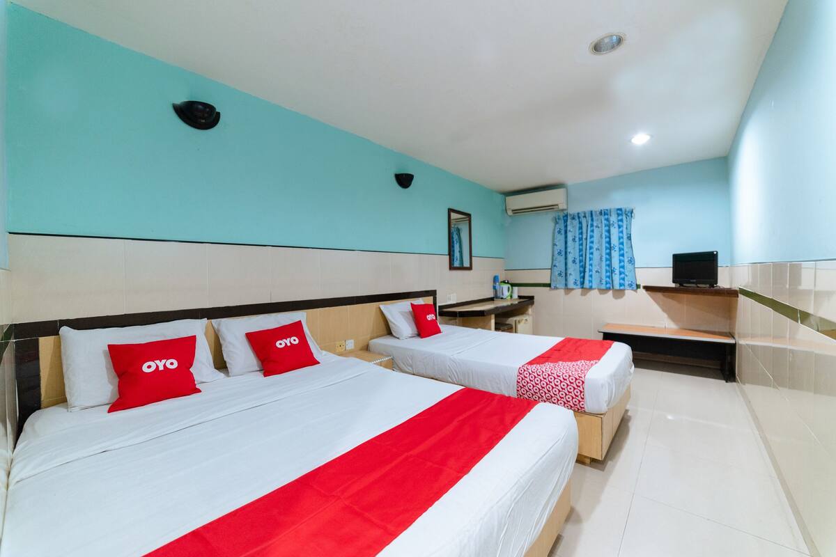 A hotel room with two beds, each with red and white bedding, a small desk, and a window with a blue curtain.