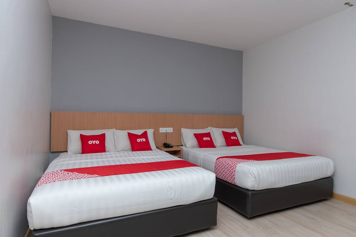 A hotel room with two beds, each with white bedding and red accents, a wooden headboard, and a neutral-colored wall.