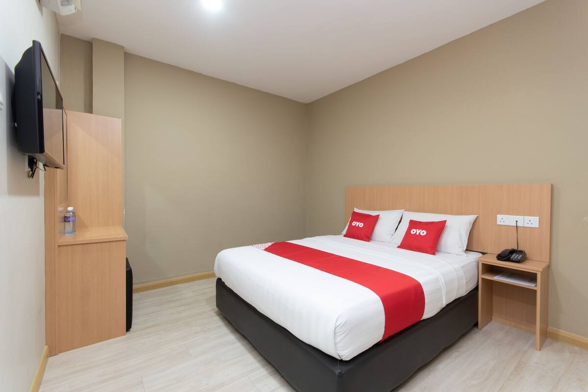 A hotel room with a bed, a TV mounted on the wall, a wooden nightstand, and a small cabinet.
