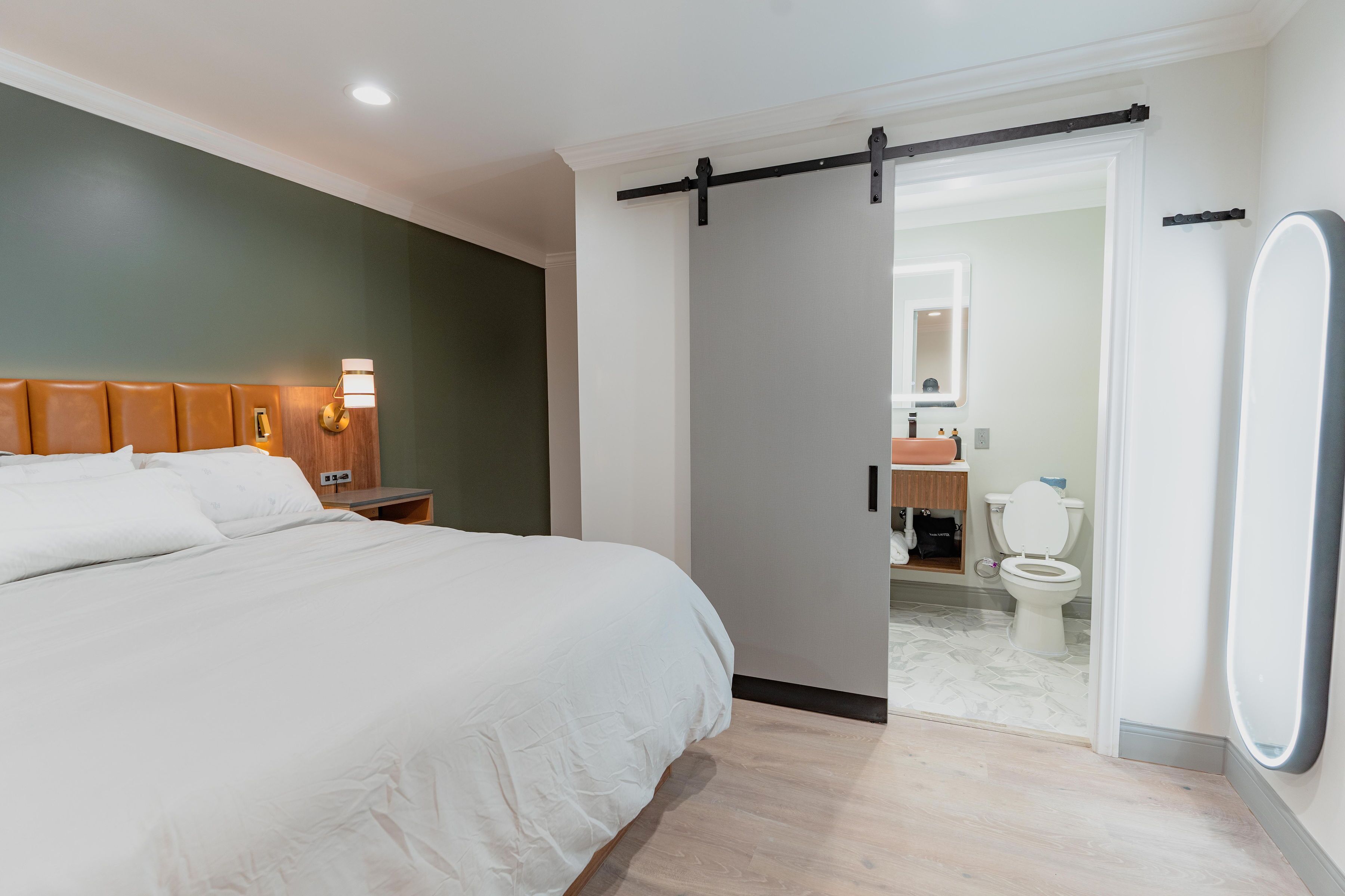 A modern hotel room with a large bed, a bedside table with a lamp, a sliding door leading to a bathroom, and a large mirror on the wall.