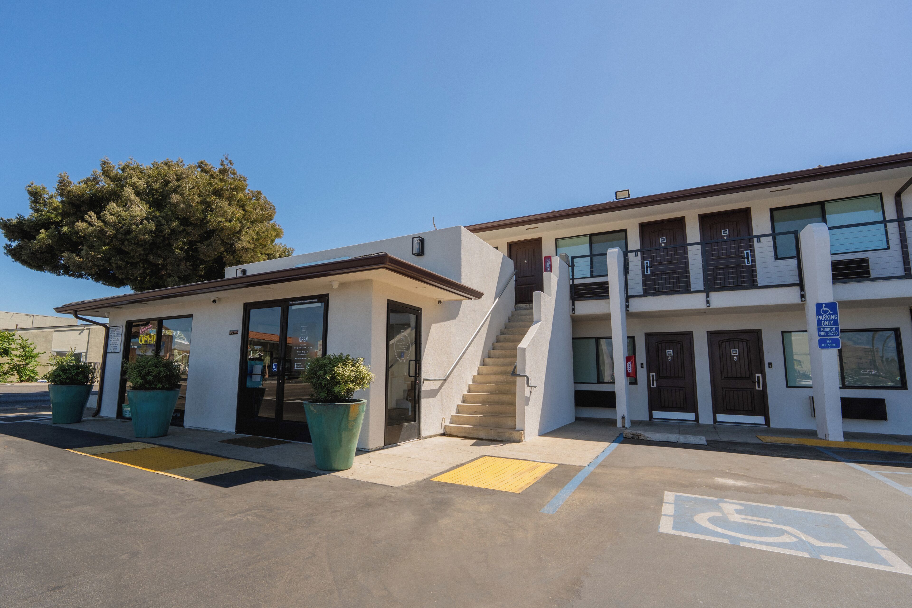 Un bâtiment doté d’un escalier menant à un étage supérieur, d’un espace de stationnement comprenant une place réservée aux personnes handicapées, et d’un grand arbre à l’avant.