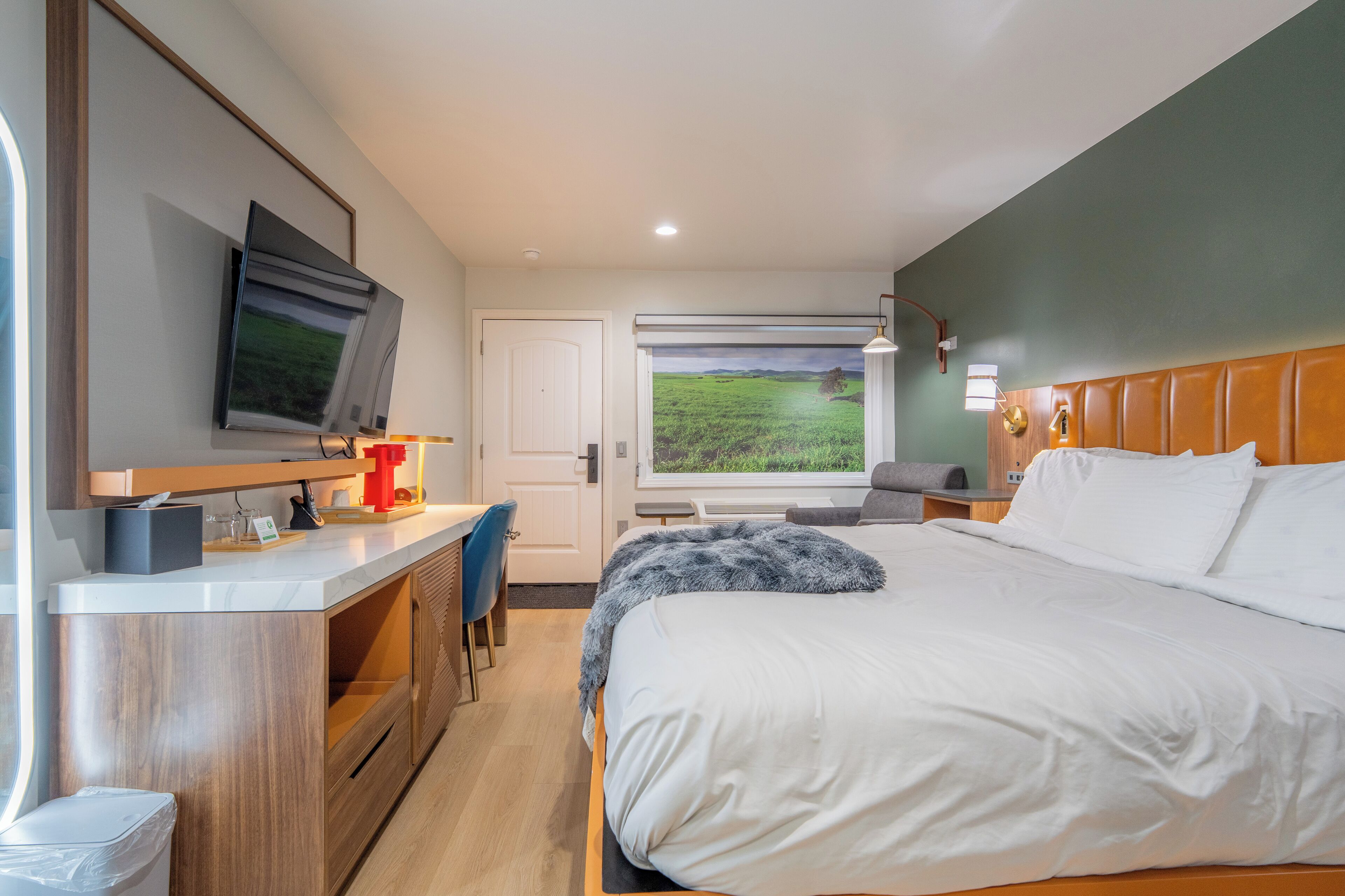A modern hotel room with a large bed, a desk, a TV mounted on the wall, and a window with a view of a green field.