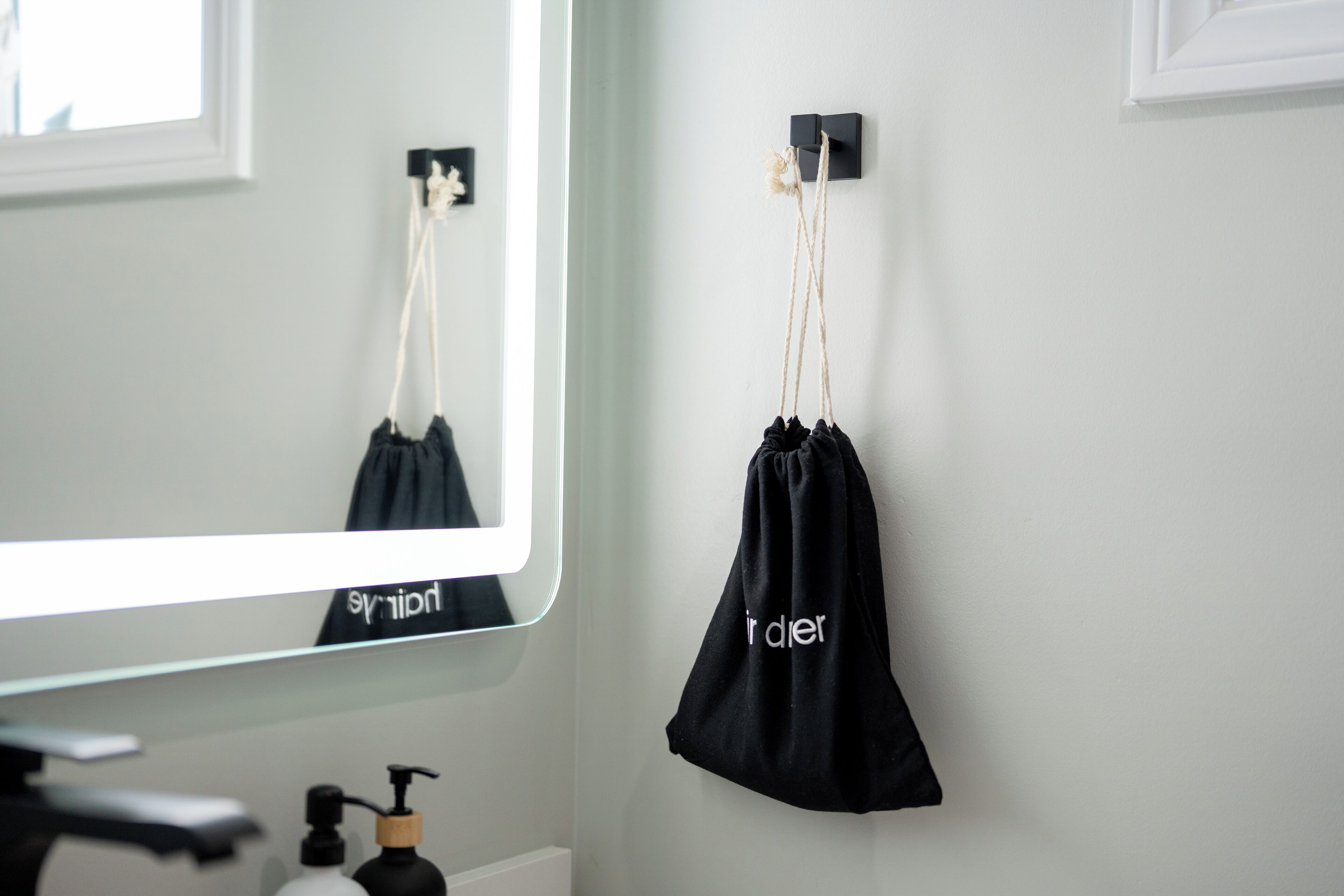 A bathroom with a mirror, a black bag hanging on a hook, and toiletries on the counter.