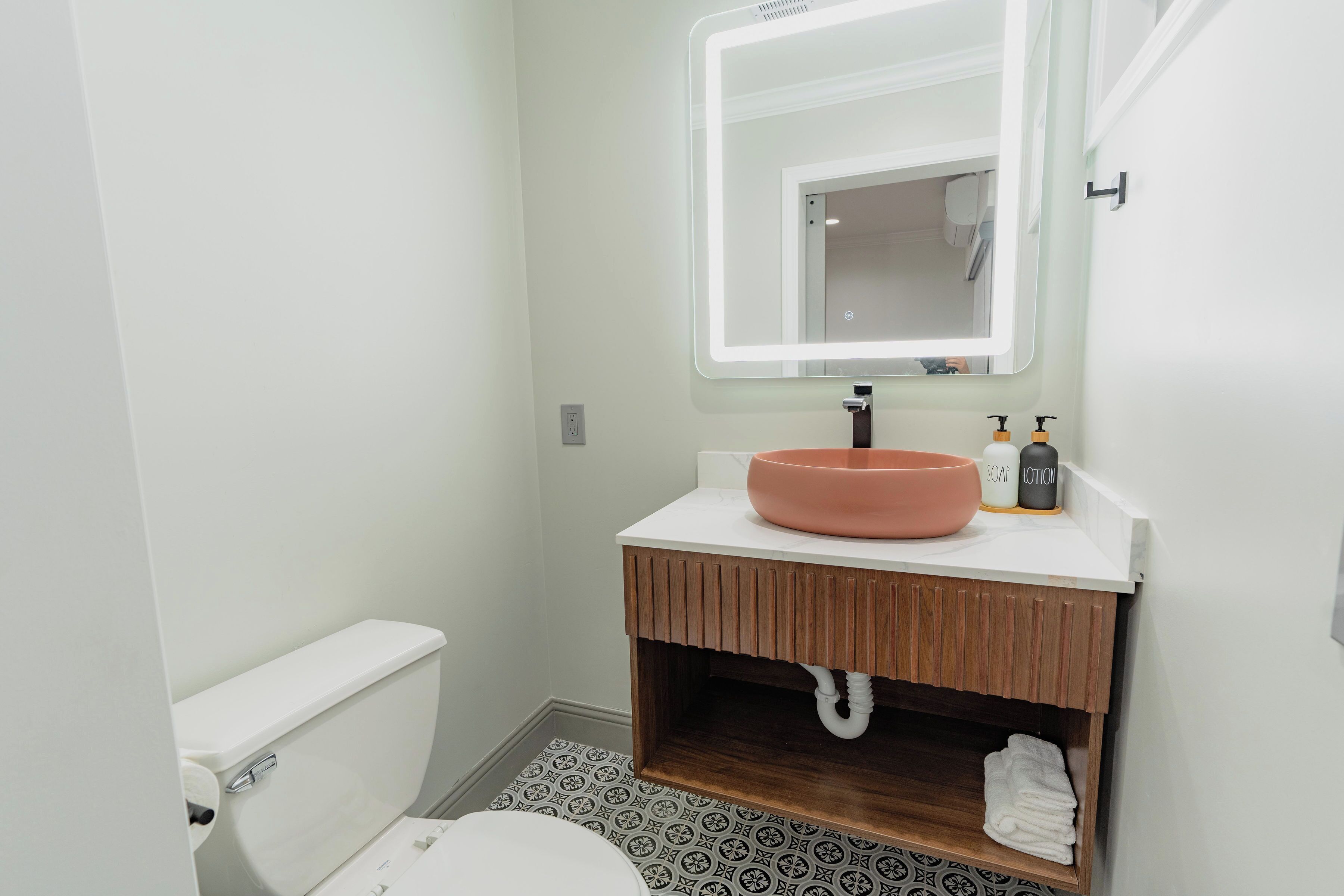 A bathroom with a white toilet, a wooden vanity with a pink basin, a mirror with built-in lighting, and a patterned floor.