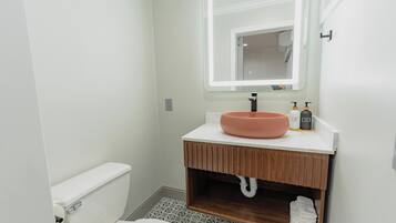 A bathroom with a white toilet, a wooden vanity with a pink basin, a mirror with built-in lighting, and a patterned floor.