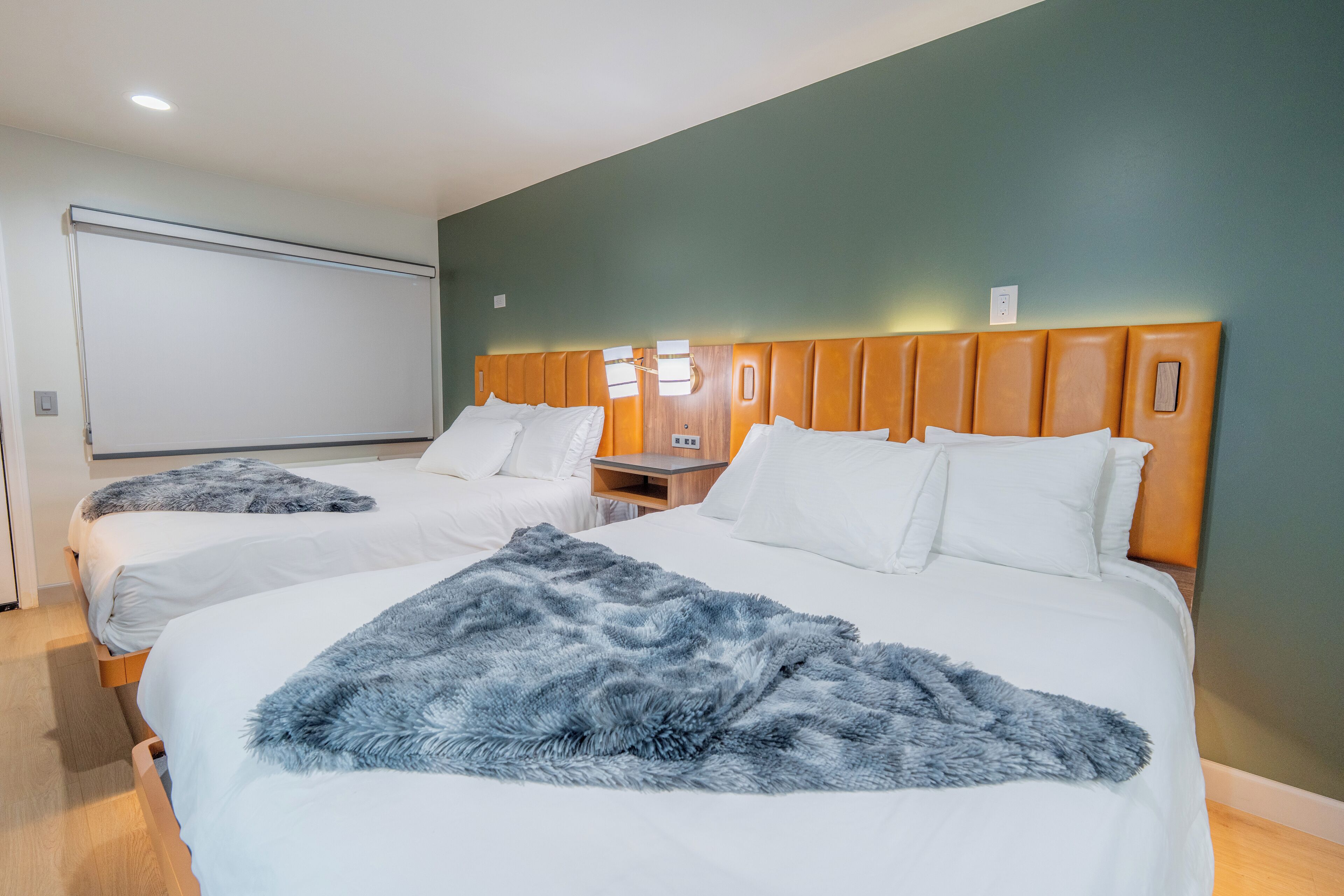 Camera d'albergo con due letti, entrambi con lenzuola bianche e una coperta azzurra soffice. La stanza ha una testiera in legno e un'ampia finestra con tende.