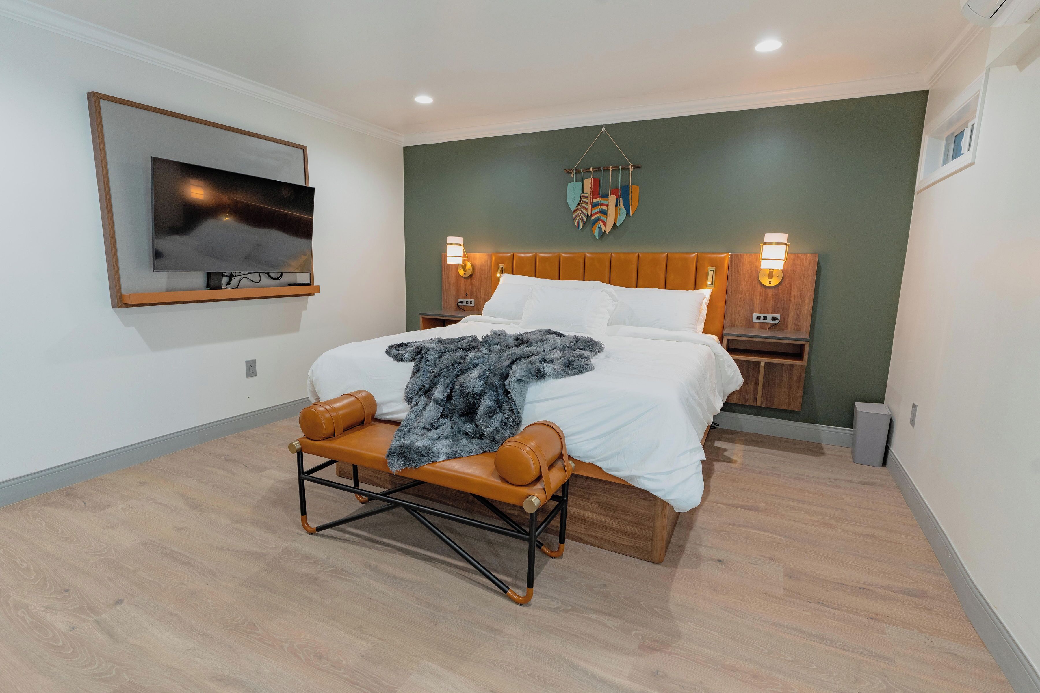 A modern bedroom with a large bed, wooden headboard, bedside lamps, a mounted TV, and a decorative wall hanging.