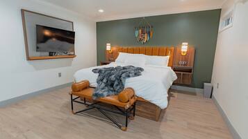 A modern bedroom with a large bed, wooden headboard, bedside lamps, a mounted TV, and a decorative wall hanging.