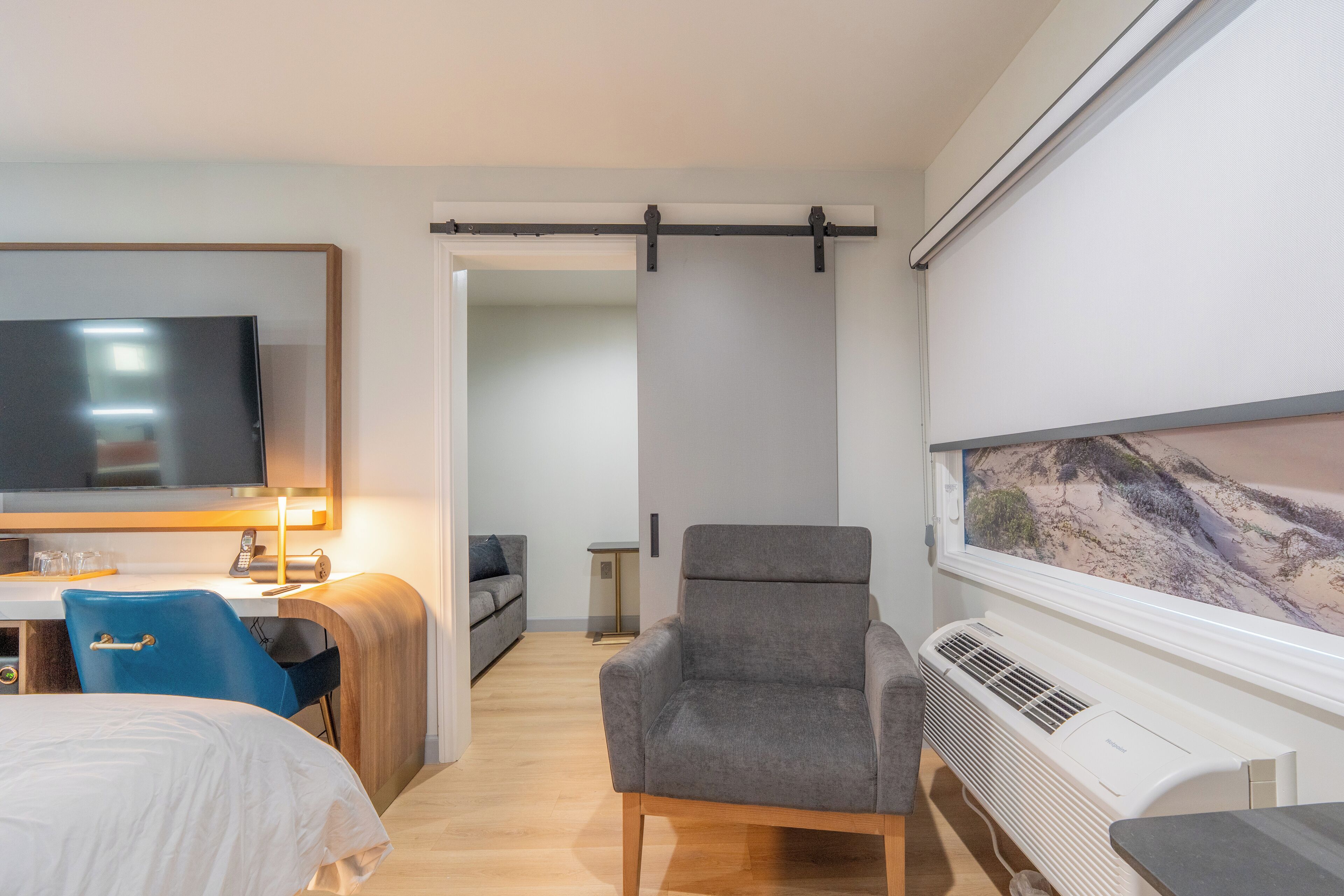 A hotel room with a bed, a desk with a computer, a chair, a TV, a window with a view, and an air conditioning unit.
