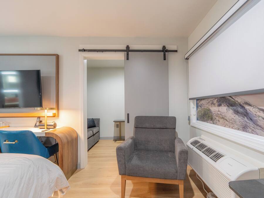 Ein Hotelzimmer mit Bett, Schreibtisch mit Computer, Stuhl, Fernseher, Fenster mit Aussicht und Klimaanlage.