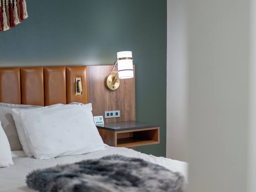 Una cama bien hecha con un edredón mullido, un cabecero de madera, una mesita de noche con una lámpara y una lámpara de pared.