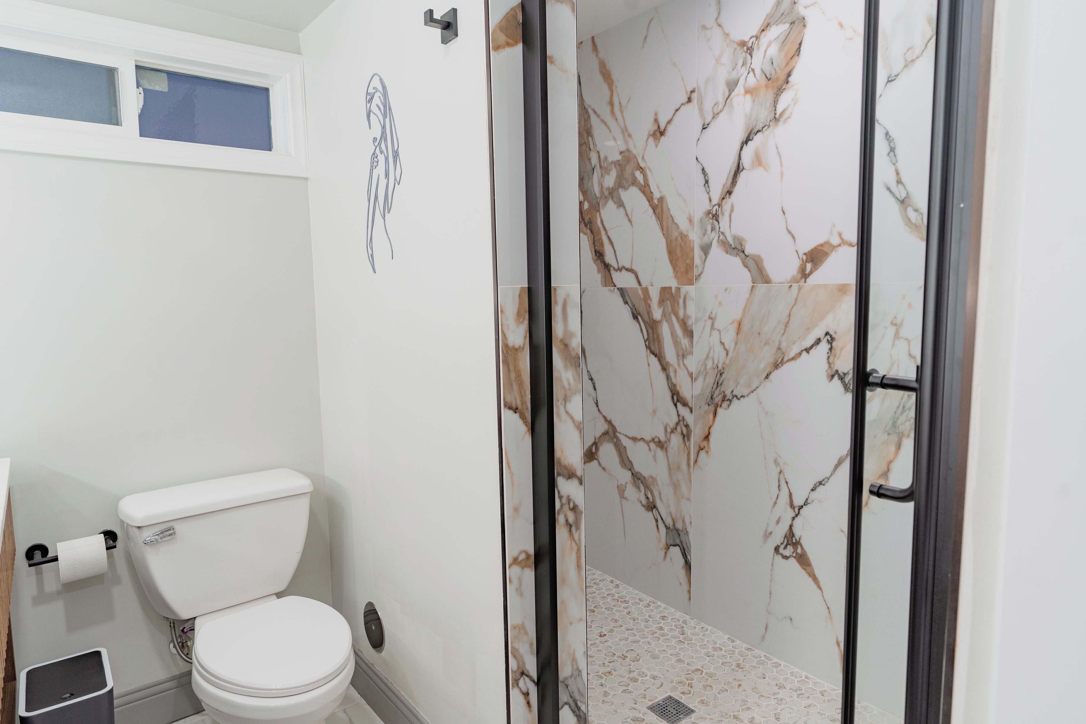 Une salle de bain avec des toilettes, une douche aux murs en marbre et une petite poubelle.