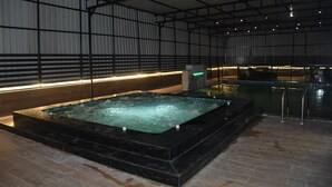 An indoor swimming pool with a black tiled edge, surrounded by a high ceiling with metal beams, and a tiled floor. - VEENA REGENCY (Chas)