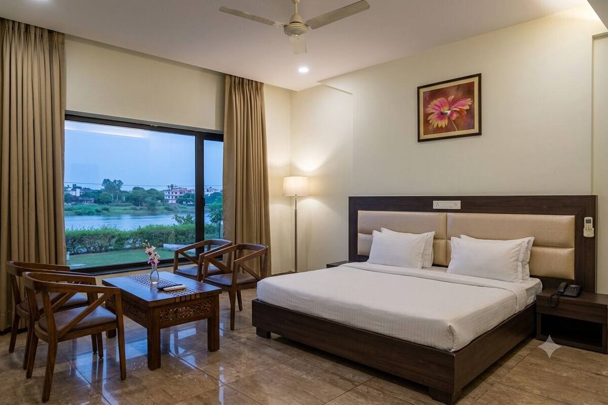 A hotel room with a bed, a desk with chairs, a lamp, a nightstand, and a view of a body of water.
