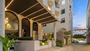 Ein modernes Gebäude mit geschwungener Fassade, einer Außenbestuhlung mit Pflanzen und einem geparkten Auto im Hintergrund.
