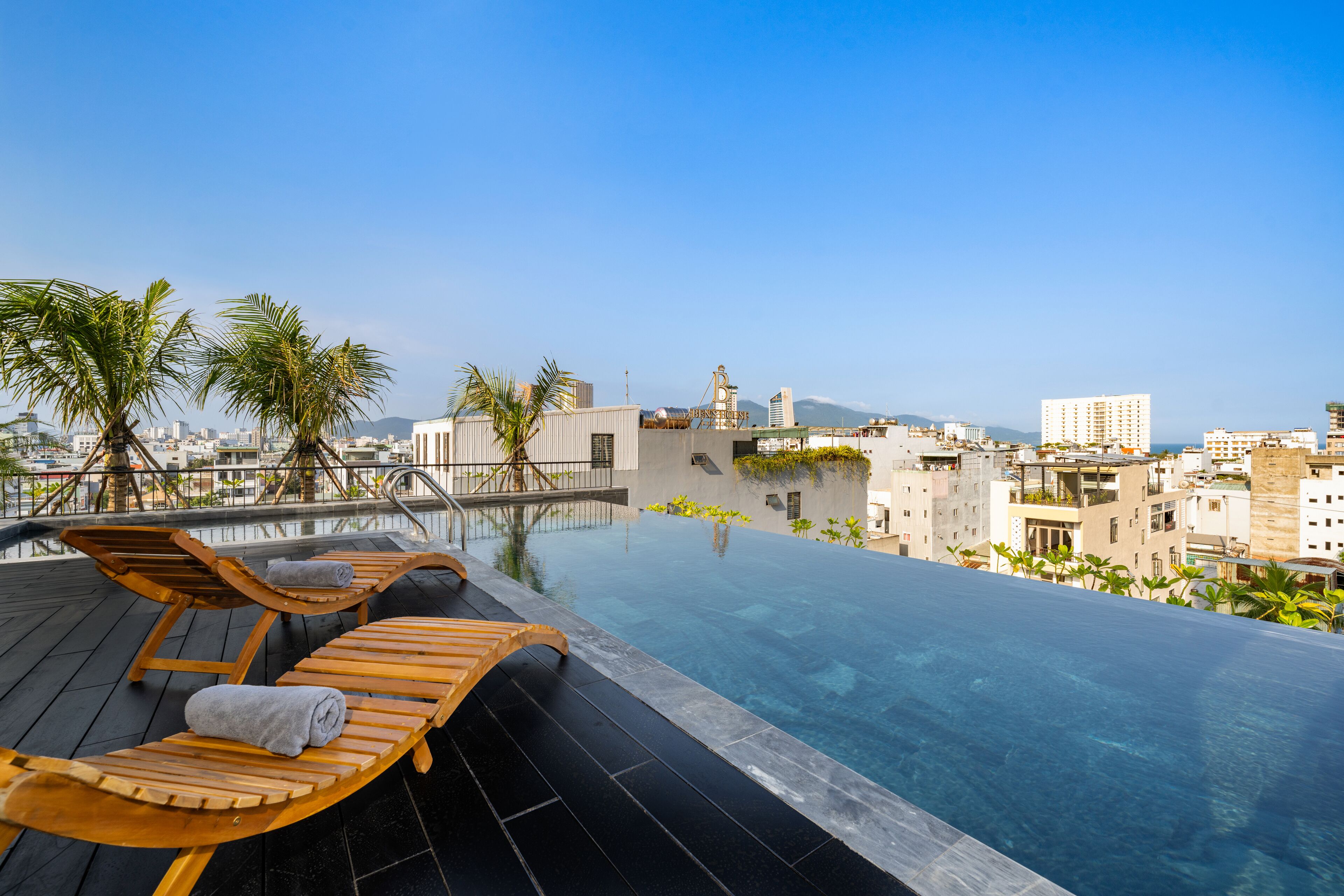 A rooftop infinity pool with wooden loungers and towels, surrounded by palm trees and buildings.