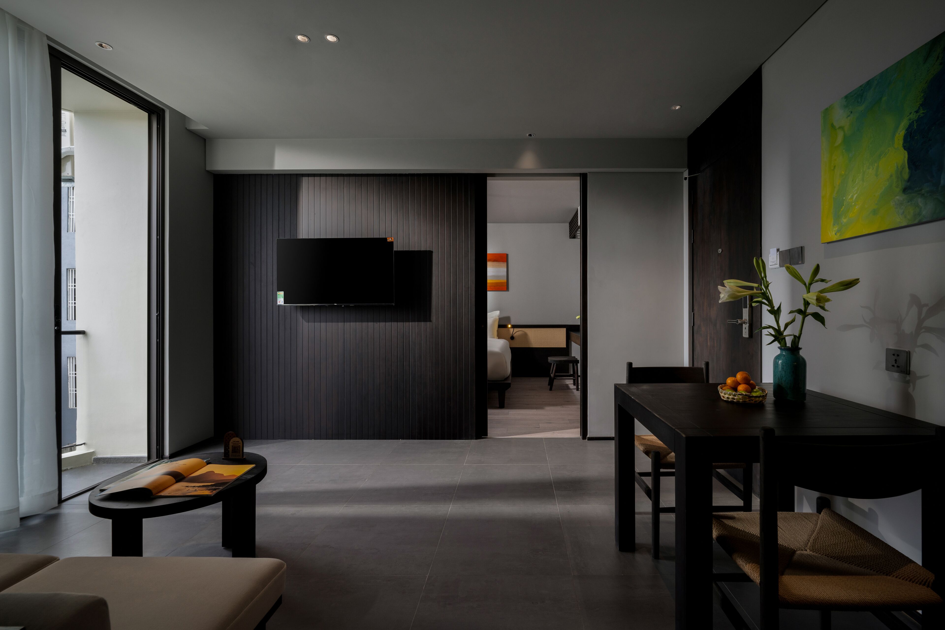 A modern living room with a flat-screen TV mounted on a dark wall, a dining area with a wooden table and chairs, and a large window providing natural light.