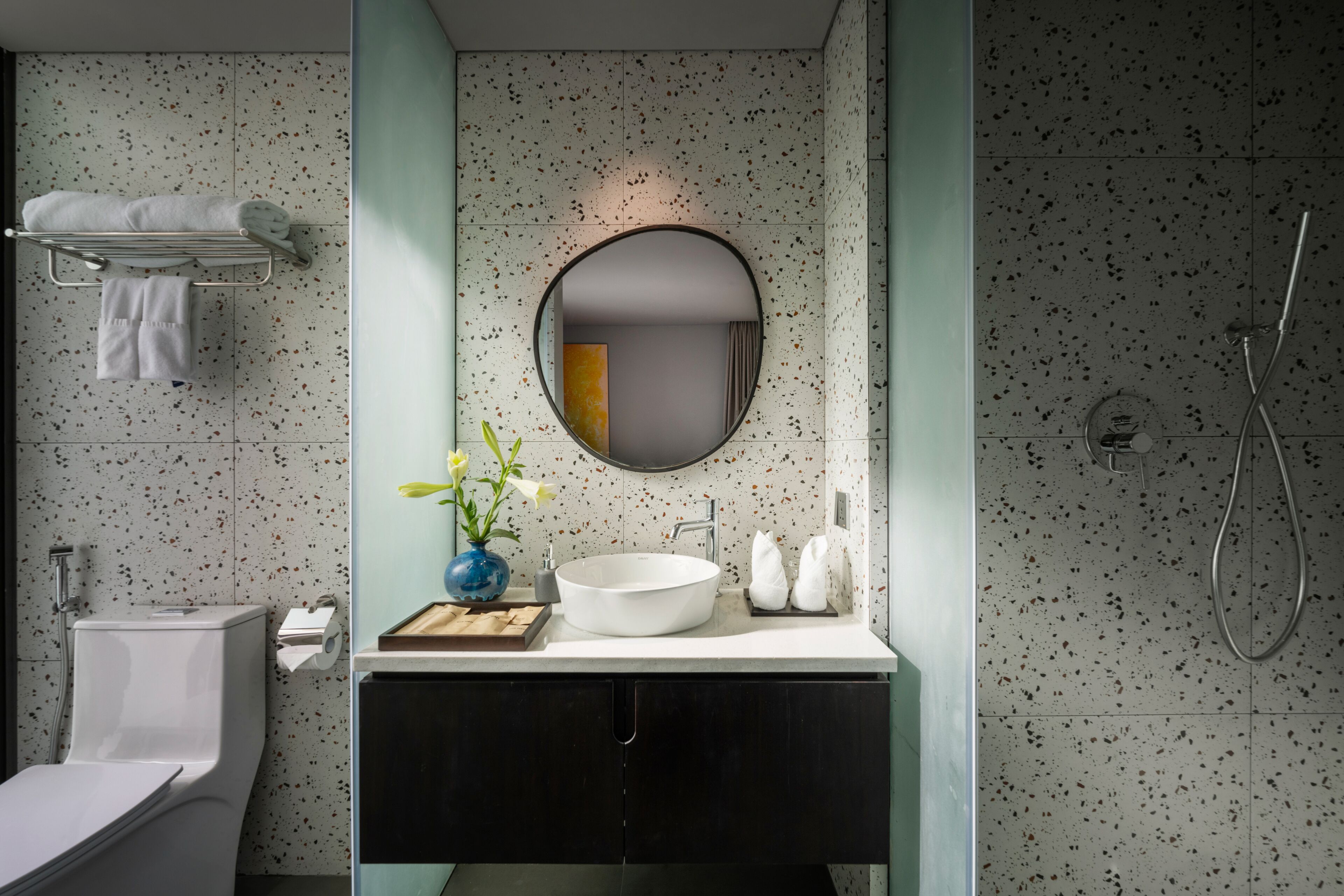 A modern bathroom with a freestanding toilet, a white sink with a round mirror, and a shower area.