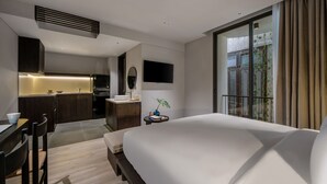 Une chambre d’hôtel moderne avec un grand lit, un bureau avec une chaise, une petite table avec une plante, un téléviseur fixé au mur et un coin cuisine à l’arrière-plan.